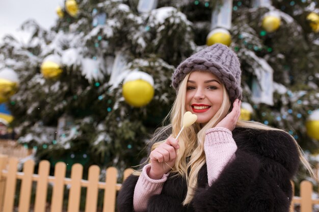 Cool blonde woman holding tasty chocolate candy against Christmas tree