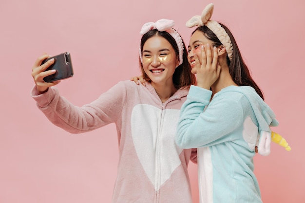 Cool brunette Asian girls in pajamas headbands and with eyepatches take selfie and sincerely smile on pink background