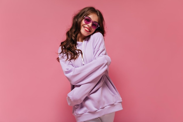 Foto cool brunette meisje in paarse hoodie en fuchsia kleur zonnebril glimlacht oprecht vrij krullend vrouw glimlacht breed op roze geïsoleerde achtergrond