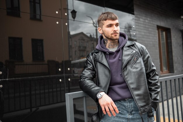 Cool brutal hipster fashion man model with a tattoo on his neck wearing trendy casual clothes with a black leather jacket and hoodie stands near a black modern building on the street