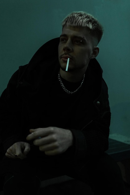 Cool brutal man in black clothes sits outside and smokes a cigarette in the night city