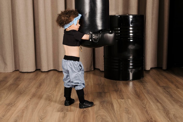 Photo cool cute little girl with afro hair and a blue bandana on the forehead wearing boxing gloves and playing with punching bag