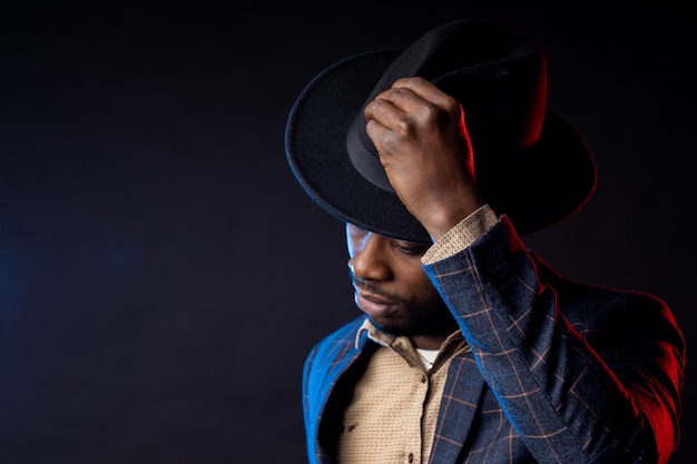 Photo cool dark skinned man in stylish suit, touching his black hat, looking down. closeup portrait of fashionable african american male model posing on dark background with backlight.