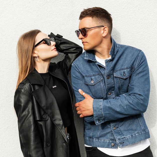 Cool fashion stylish couple man and woman with fashionable sunglasses in trendy casual denim clothes pose near a wall in the city