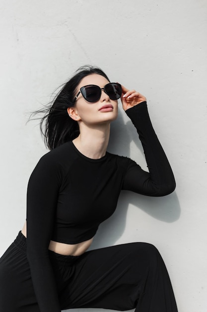 Photo cool fashion urban trendy pretty woman in fashionable black sports outfit wears a stylish black sunglasses and poses near a gray wall on the street