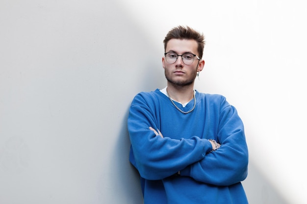 Cool fashion young hipster man with glasses in blue sweatshirt stands near a white wall on the street