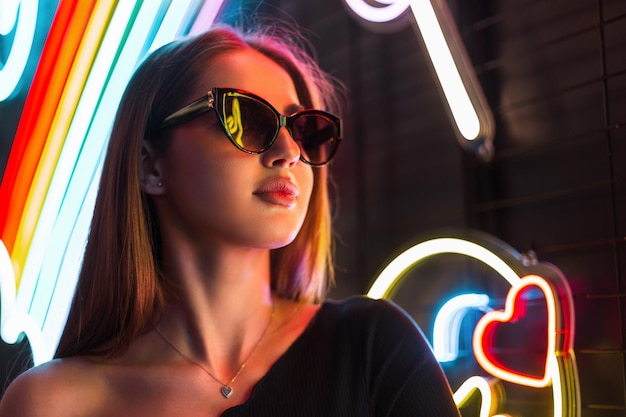 Photo cool female trendy portrait of a beautiful woman with stylish sunglasses wearing fashion black outfit on a dark background with neon lights