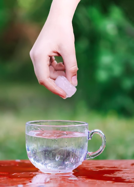 Acqua fresca fresca e cubetto di ghiaccio in mano sopra la tazza di vetro trasparente nel tavolo all'aperto nelle giornate estive