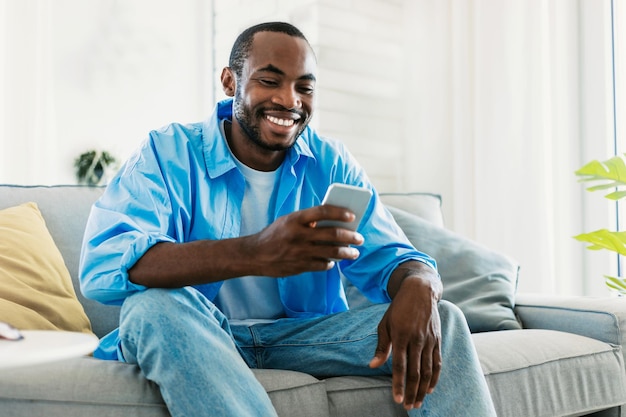 Cool gadget and app concept portrait of happy black man using\
mobile phone watching video sitting on sofa copy space