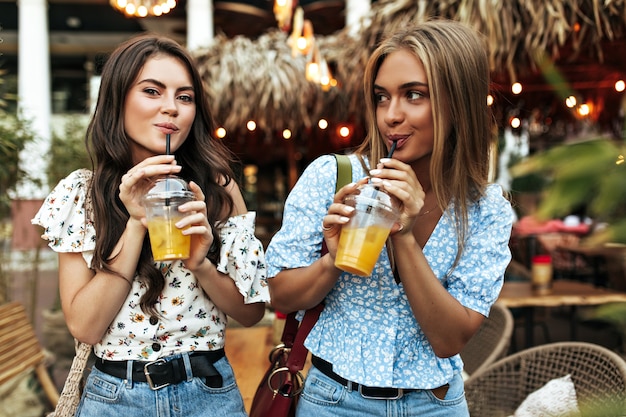 Cool gebruinde brunette en blonde vrouwen in spijkerbroeken en stijlvolle blouses met bloemen lopen goedgehumeurd naar buiten en drinken limonade