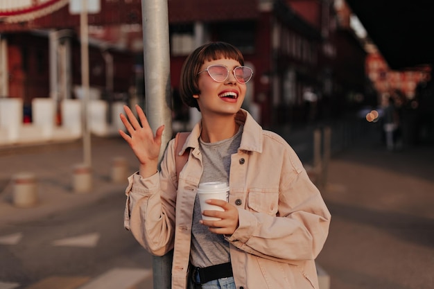 Cool girl in beige jacket walks outdoors Happy brunette woman in sunglasses holding cup of coffee and waving hand outside