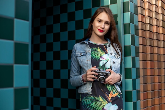 Cool girl model with retro film camera wearing a denim jacket, dark hair outdoors over city wall in a cage background. Having fun in the city with camera, travel photo of photographer.