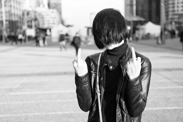 Cool girl with stylish short brunette hair in black leather jacket show middle finger outdoor on street background