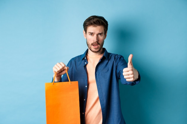 Cool guy showing thumb up and holding orange shopping bag, standing on blue background.