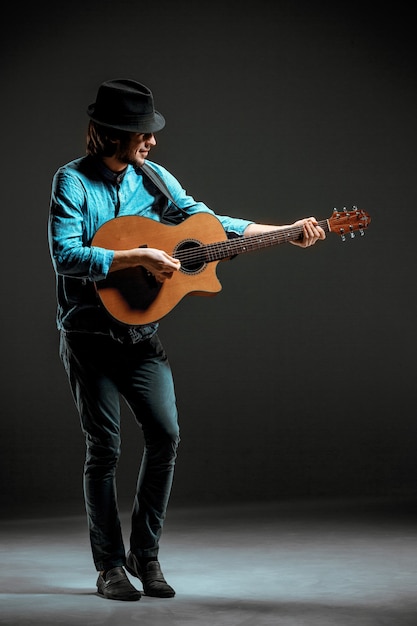 Ragazzo figo con cappello in piedi con la chitarra su sfondo scuro studio