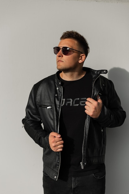 Cool handsome hipster guy in a fancy black leather jacket Tshirt and sunglasses near a gray concrete wall