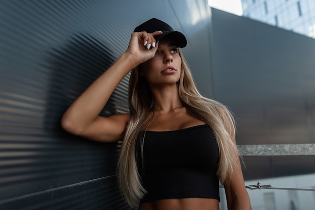 Cool hipster blonde woman with fashion black cap in black t-shirt top posing near dark modern wall in city