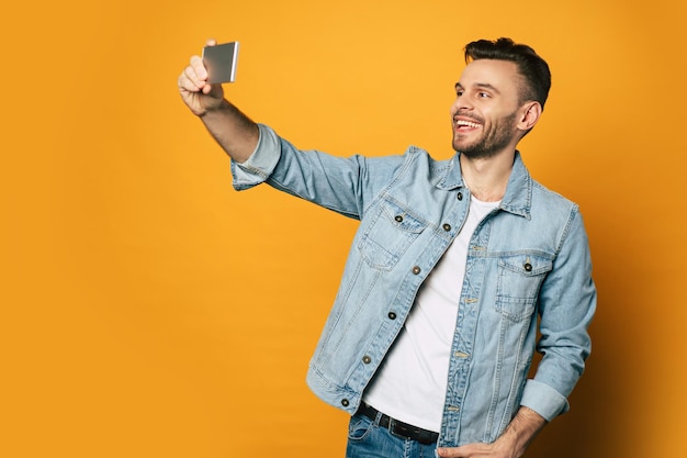 Cool man in denim blue jacket and white T-shirt with dark brown straight hair and perfect white smile is using a camera on his phone for talking to friends