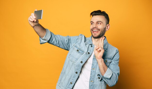 Cool man in denim blue jacket and white T-shirt with dark brown straight hair and perfect white smile is using a camera on his phone for talking to friends