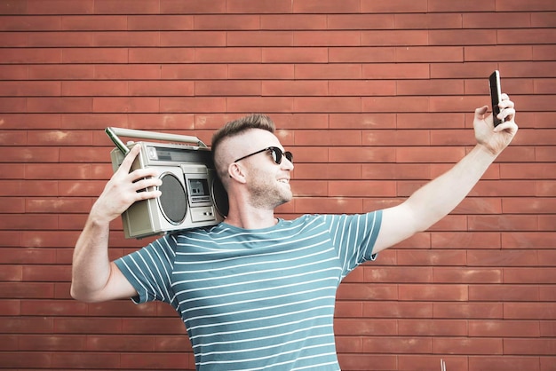 Cool man taking a selfie while listening music with 80\'s\
boombox stereo outdoors in the street guy having fun dancing with\
vintage radio joyful youth lifestyle tech concept red wall\
back