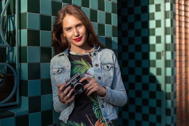 Cool meisjesmodel met retro filmcamera met een spijkerjasje, donker haar buiten over de stadsmuur op een kooiachtergrond. Plezier hebben in de stad met camera, reisfoto van fotograaf.