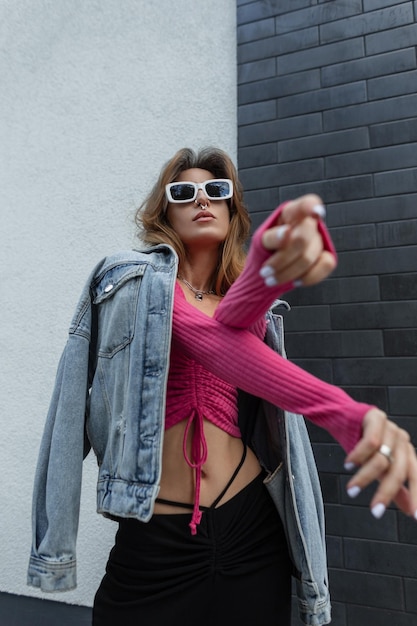 Cool mode jonge roodharige vrouw model hipster met witte stijlvolle zonnebril in een trendy roze sweatshirt top met denim jasje en rok dansen in de buurt van een bakstenen zwarte muur op straat