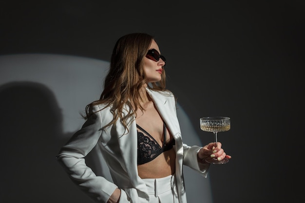 Cool mode schoonheid sexy vrouw met stijlvolle zonnebril in een mode wit pak met een blazer en zwarte kanten beha met een vintage glas champagne op een donkere achtergrond in de studio