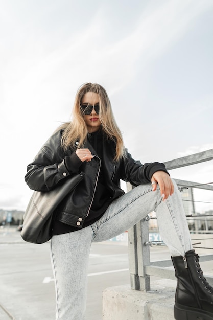Cool modieuze jonge vrouw model hipster in mode lente outfit met lederen rock jas hoodie zwarte tas spijkerbroek en leren schoenen laarzen poses in de stad Urban vrouwelijke stijl look kleding