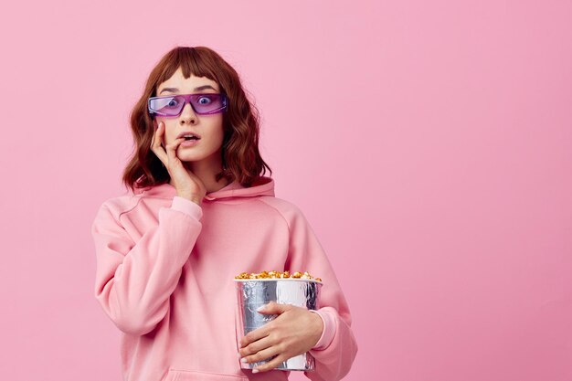 Cool movie Confused cute redhead lady in pink hoodie sunglasses with popcorn posing isolated on over pink studio background show copy space Banner Fashion Cinema concept Entertainment offer