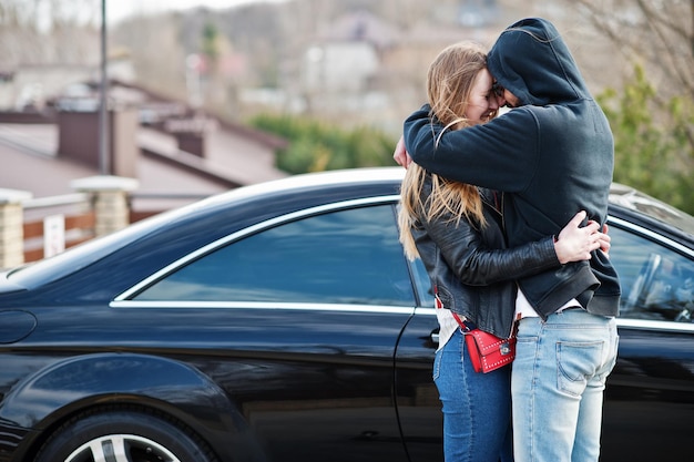 Cool multiracial couple hugs together against black elegant sport car