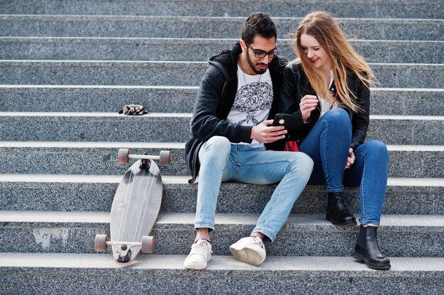 Cool multiracial couple sitting on stairs with longboard and look in mobile phone.