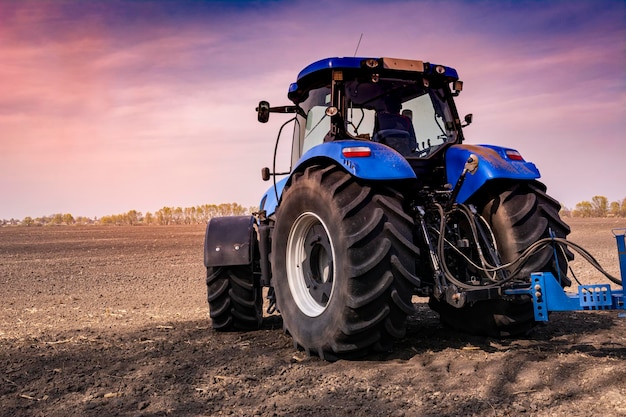 A cool new blue tractor in a field at sunset processes the field. Agricultural machinery works in the field. Purple sky on a background.