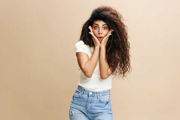 Cool offer excited pretty latin female in white tshirt with\
afro open mouth recline on hands smile at camera say wow stay\
isolated over beige background copy space free place