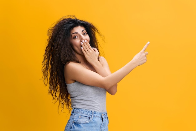 Cool offer shocked pretty latin female with afro point finger\
up close mouth with hand show empty place stay isolated over yellow\
background gossip concept copy space free place