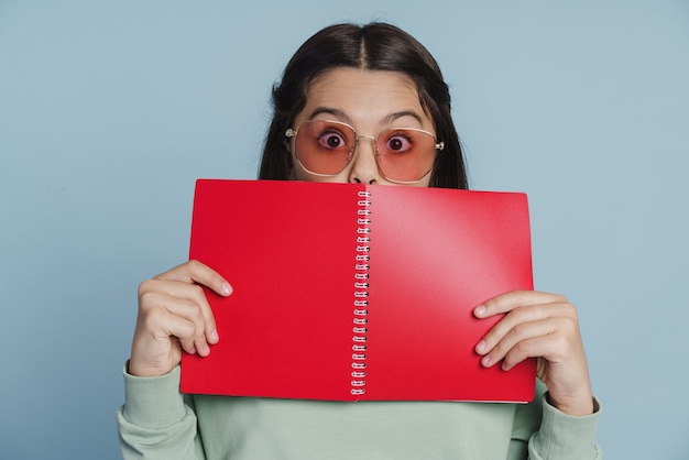 Cool, positive teen girl covers her face with a notebook. Little girl in sunglasses on a blue background