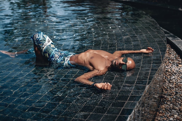 Cool and relaxed Arabic shirtless male in turquoise sarong lying in the pool in a sunny day; vacation concept.