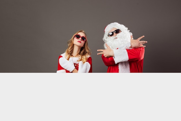 Photo cool santa claus and young beautiful mrs. claus in sunglasses stand behind a white canvas