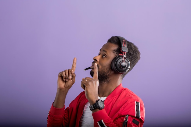 Cool serious african american man dressed in wearing red windbreaker jacket, dancing, listening to music in modern headphones on purple studio background.