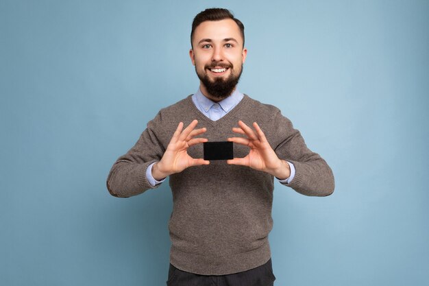 Cool smiling brunette bearded man wearing grey sweater and blue shirt isolated on background wall holding credit card looking at camera.