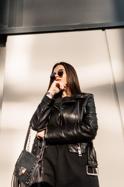 Cool stijlvolle mooie hipster vrouw in fashion zwarte rock kleding met leren jas en vintage ronde zonnebril in de buurt van de muur in de stad