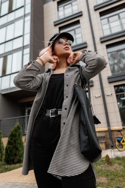 Cool stylish beautiful woman with glasses and a vintage cap in casual fashion jeans clothes with a bag walks in the city near the building