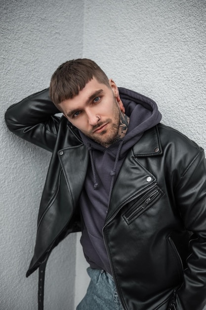cool stylish brutal handsome man model with hair and beard with a tattoo on his neck in fashionable rock clothes with leather jacket and hoodie stands and poses near the white wall in the city