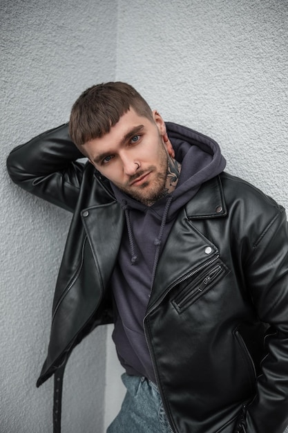 Cool stylish brutal handsome man model with hair and beard with a tattoo on his neck in fashionable rock clothes with leather jacket and hoodie stands and poses near the white wall in the city