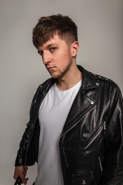 cool stylish man in a fashionable leather black jacket and a white t-shirt on a gray background in the studio