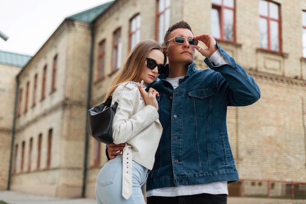 Cool stylish models couple in trendy casual spring clothes with fashion vintage sunglasses are walking in the city near the buildings Fashion urban style man's and woman's look outfit