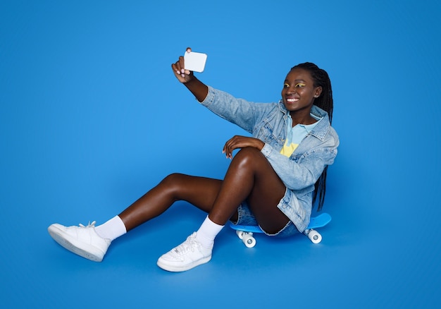 Cool stylish young black woman sitting on skateboard taking selfie