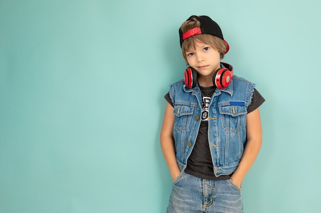 A cool tenager boy in denim jaket and shorts, red earphones, black cap, stands in front of the camera and keeps his hands in pockets, isolated on blue background