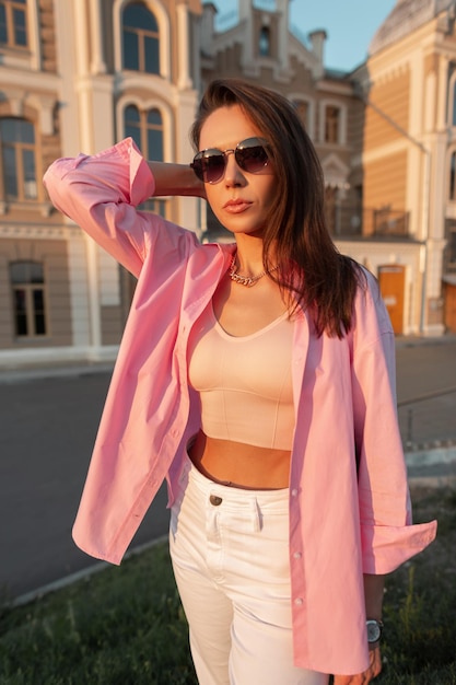 Cool trendy beautiful girl with fashion sunglasses in pink shirt with tank top walks in the city Urban glamour female style look