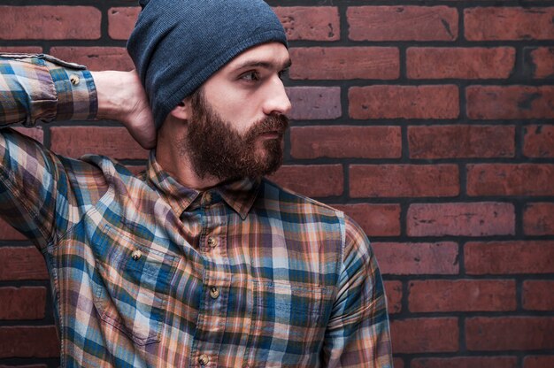 Photo cool and trendy. confident young bearded man holding hand behind head and looking away while standing against brick wall
