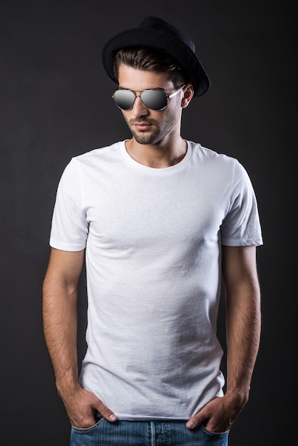 Cool and trendy. Handsome young man in sunglasses and fedora keeping hands in pockets while standing against black background